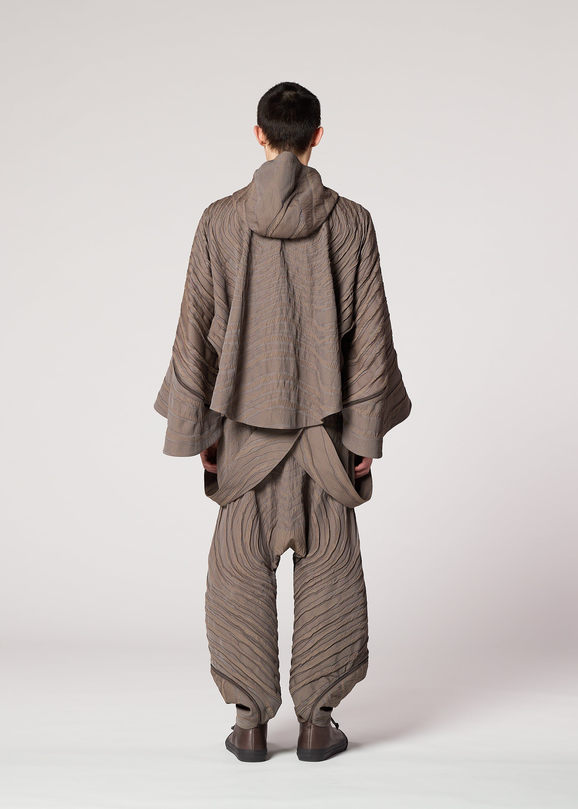 Zoomed view A person faces away, wearing IM MEN's ENGRAVE: a Shoji Kamoda-inspired taupe outfit with wide pants and a hooded, layered top. The textured design evokes three-dimensional wave patterns against a light background.
