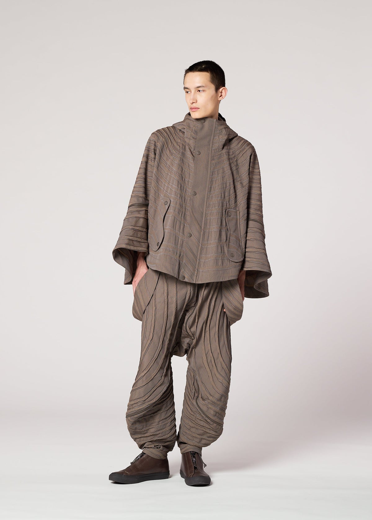 A person wears the IM MEN ENGRAVE set: a Shoji Kamoda-inspired jacquard loom jacket and wide balloon pants in taupe. The textured fabric has stitched, curved lines for a wave effect. Dark shoes complete the outfit. view 8 of 9