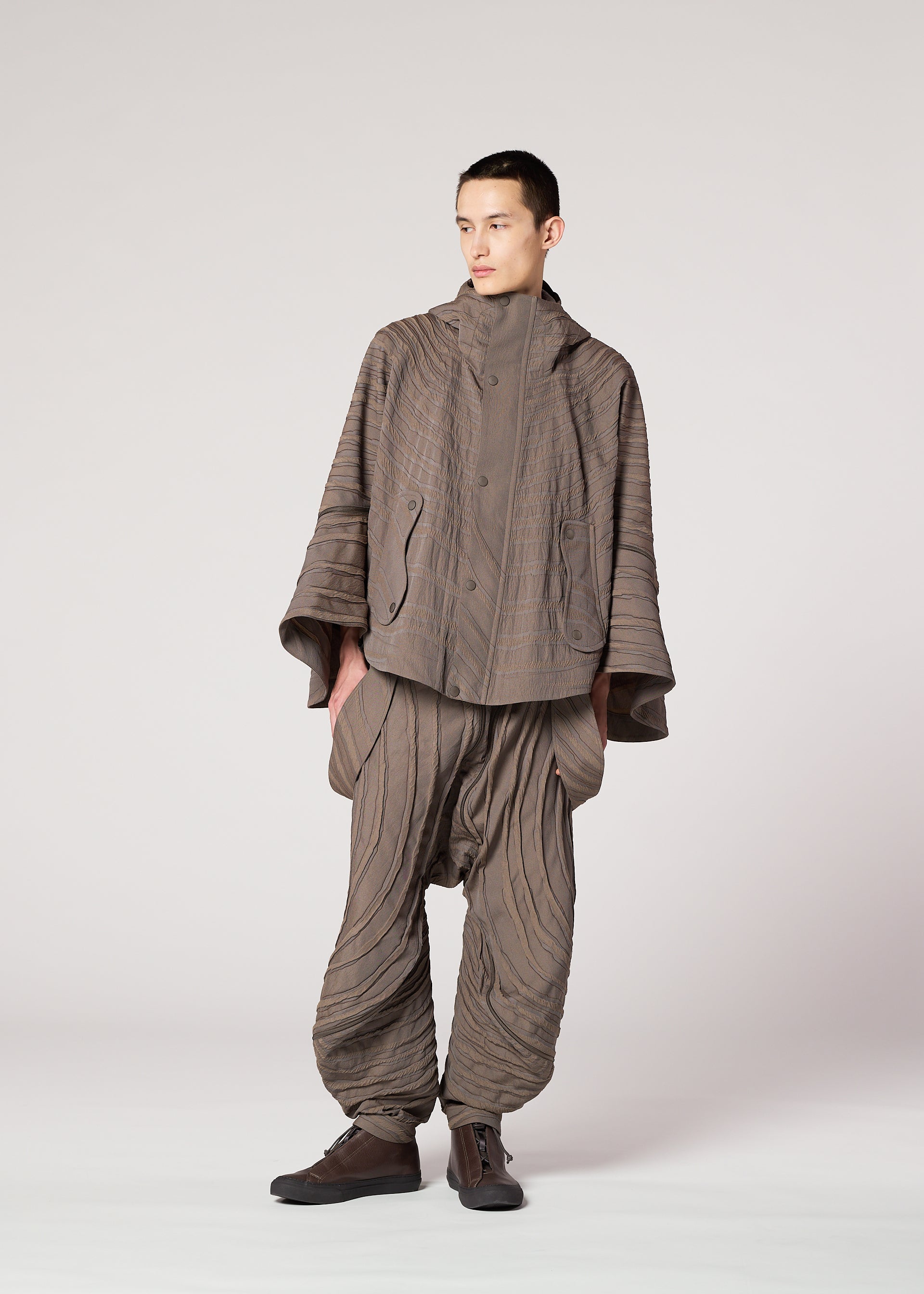 Zoomed view A person wears the IM MEN ENGRAVE set: a Shoji Kamoda-inspired jacquard loom jacket and wide balloon pants in taupe. The textured fabric has stitched, curved lines for a wave effect. Dark shoes complete the outfit.