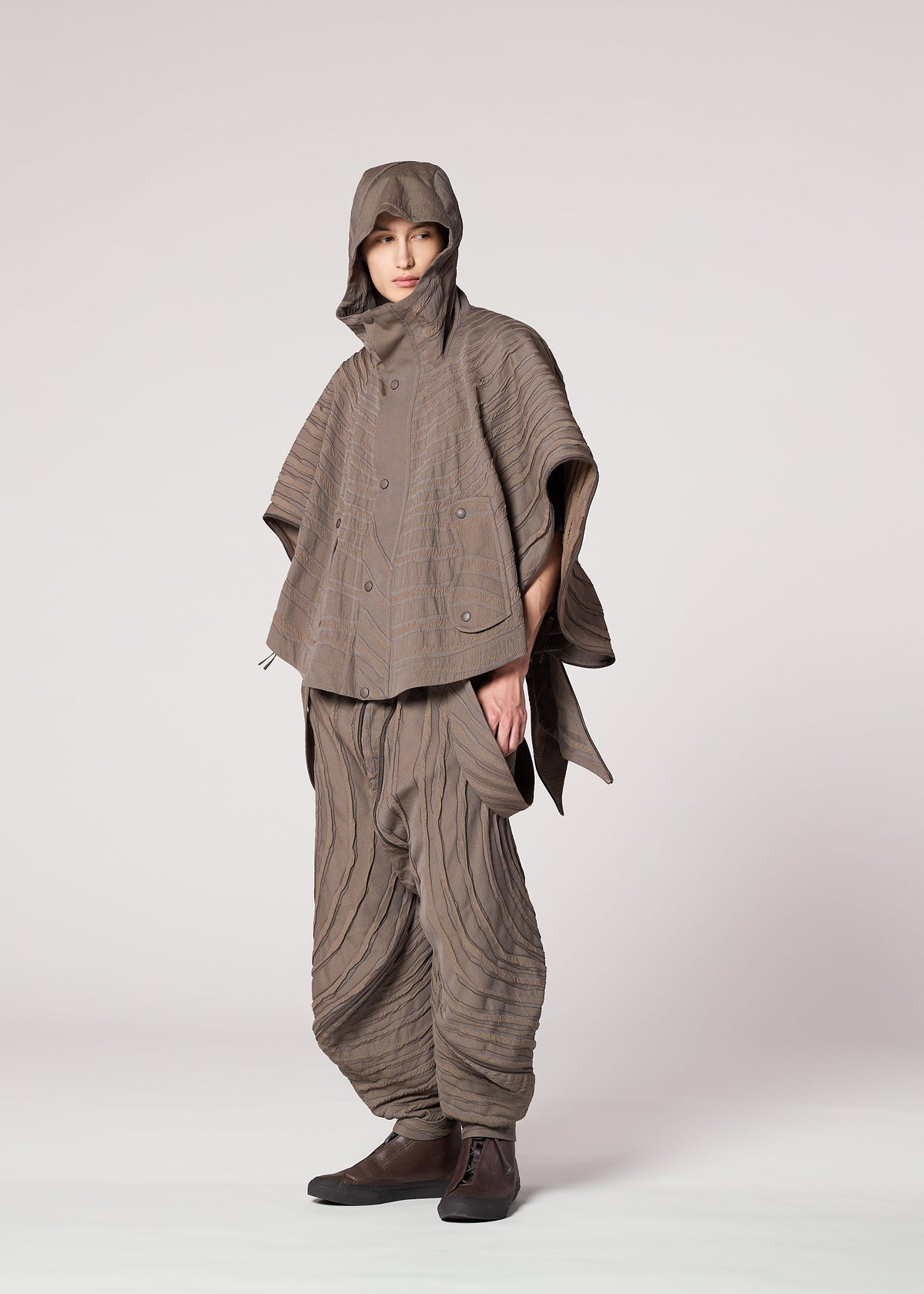 A person stands in a studio wearing IM MEN’s ENGRAVE: a taupe, Shoji Kamoda-inspired outfit with a hooded poncho and loose layered pants featuring sculpted wave patterns, dark shoes, and one hand in their pocket. view 9 of 9
