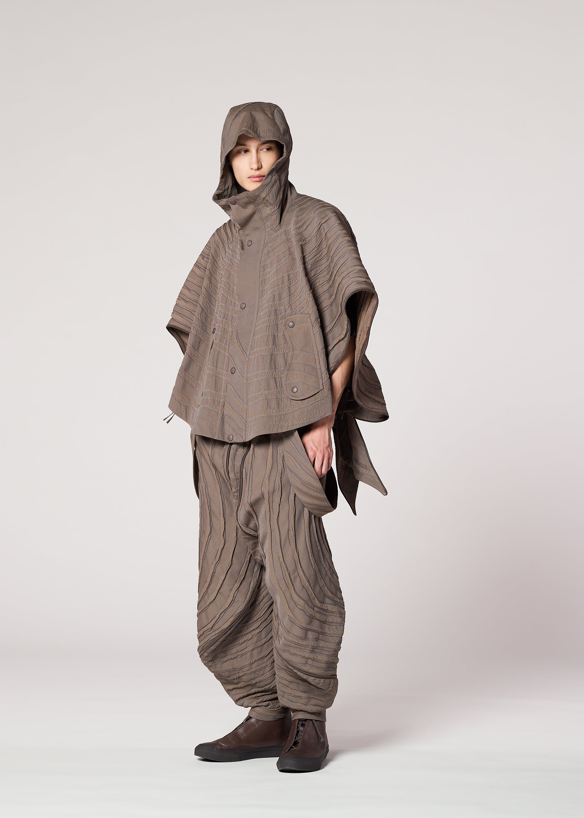 Zoomed view A person stands in a studio wearing IM MEN’s ENGRAVE: a taupe, Shoji Kamoda-inspired outfit with a hooded poncho and loose layered pants featuring sculpted wave patterns, dark shoes, and one hand in their pocket.