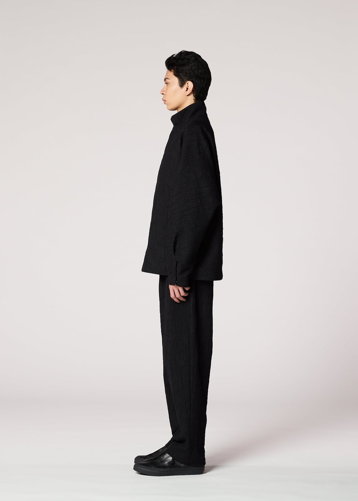 A person with short dark hair stands in profile against a plain light background, wearing IM MEN’s SHRINK JACQUARD—a loose-fitting black jacket with unique texture—paired with matching pants and black shoes. view 6 of 8