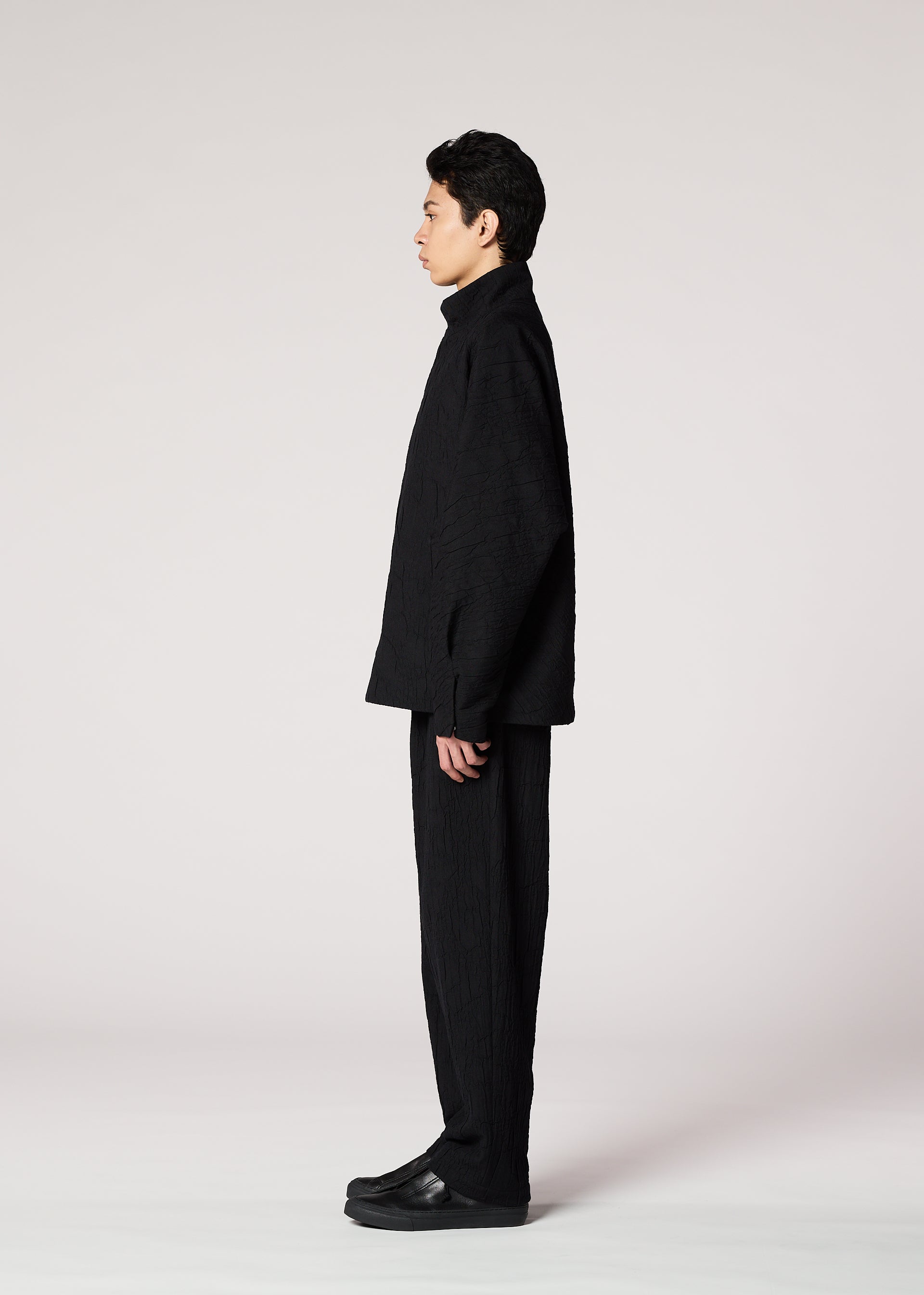 Zoomed view A person with short dark hair stands in profile against a plain light background, wearing IM MEN’s SHRINK JACQUARD—a loose-fitting black jacket with unique texture—paired with matching pants and black shoes.
