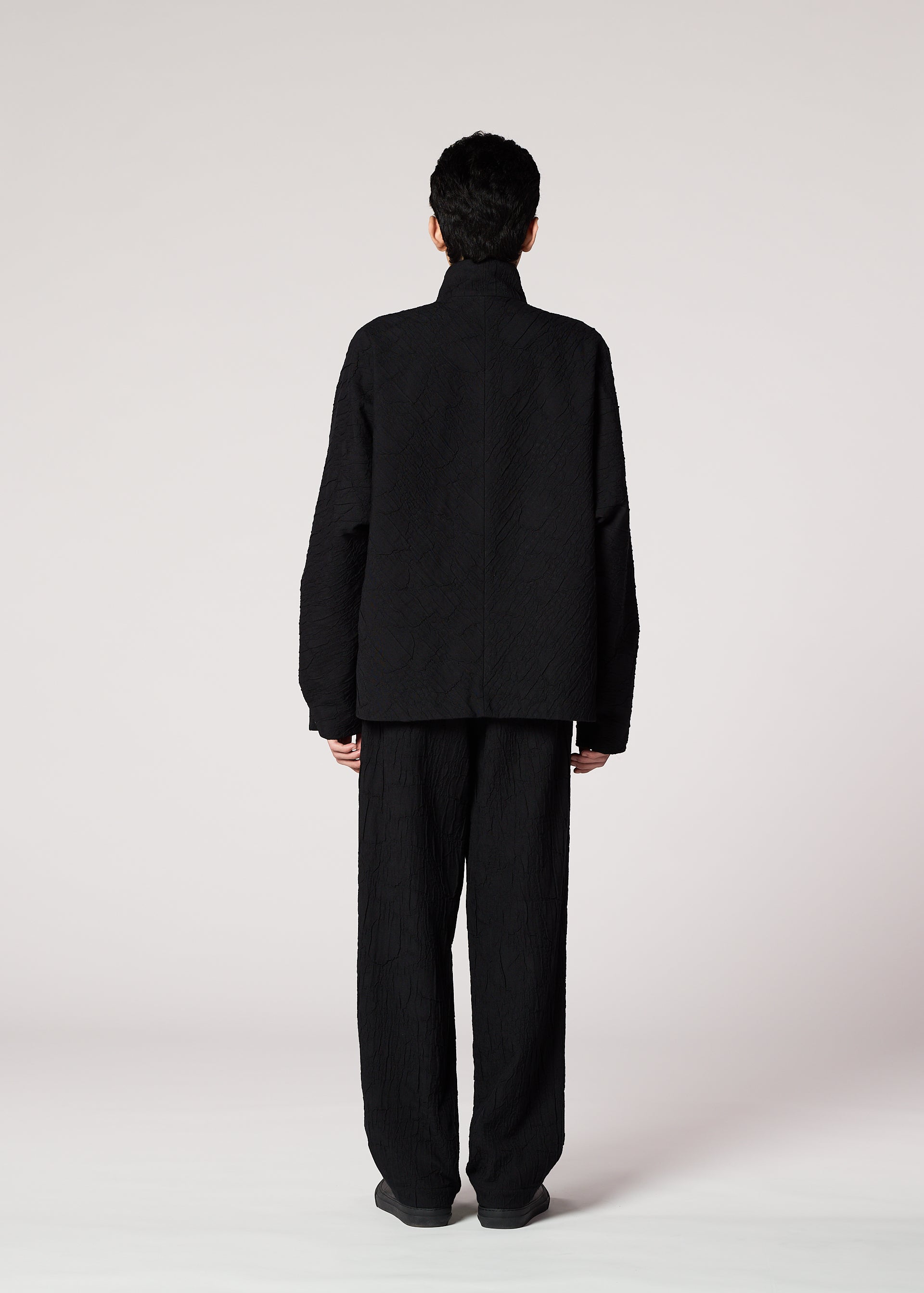 Zoomed view A person with short dark hair wears the IM MEN SHRINK JACQUARD black jacket and matching pants, standing with their back to the camera against a plain light background.