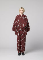 A person with short blonde hair stands against a light gray background, wearing the EARTH jacket by IM MEN, featuring a loose fit, maroon wave pattern with bold white abstract designs, paired with black shoes.