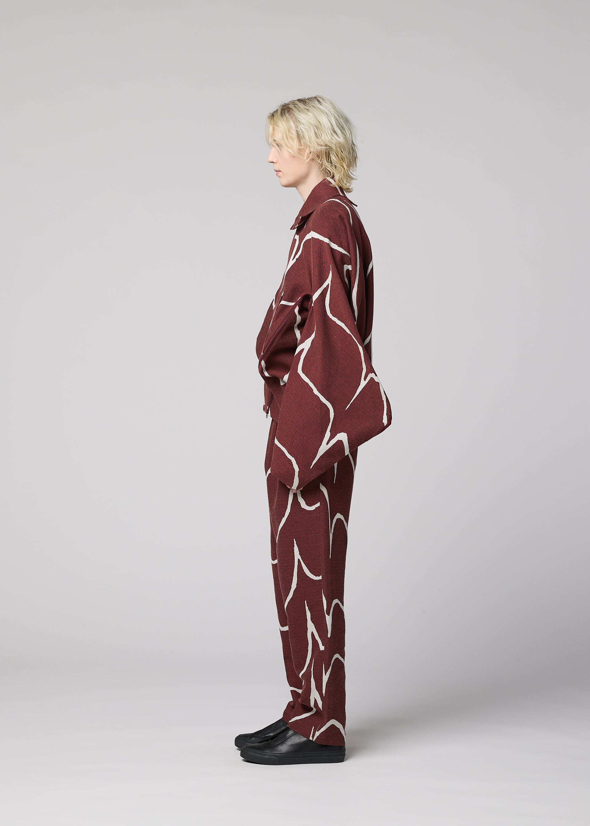Zoomed view A person with blond, wavy hair stands in profile against a plain background, wearing IM MEN’s EARTH jacket—a loose-fitting maroon piece with white abstract lines—and black shoes.