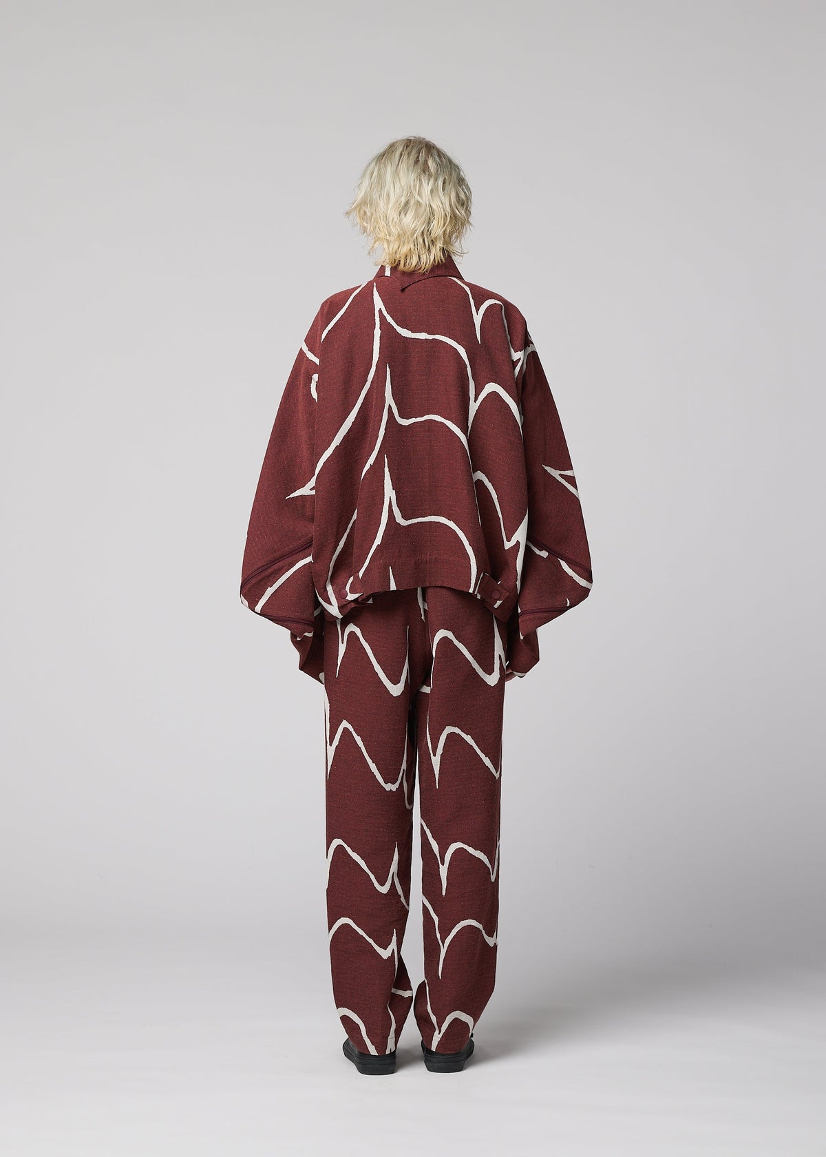A person with short, wavy blonde hair stands with their back to the camera, wearing IM MEN's EARTH— a loose maroon ceramic-inspired jacket with white abstract lines—set against a plain light gray background. view 4 of 5