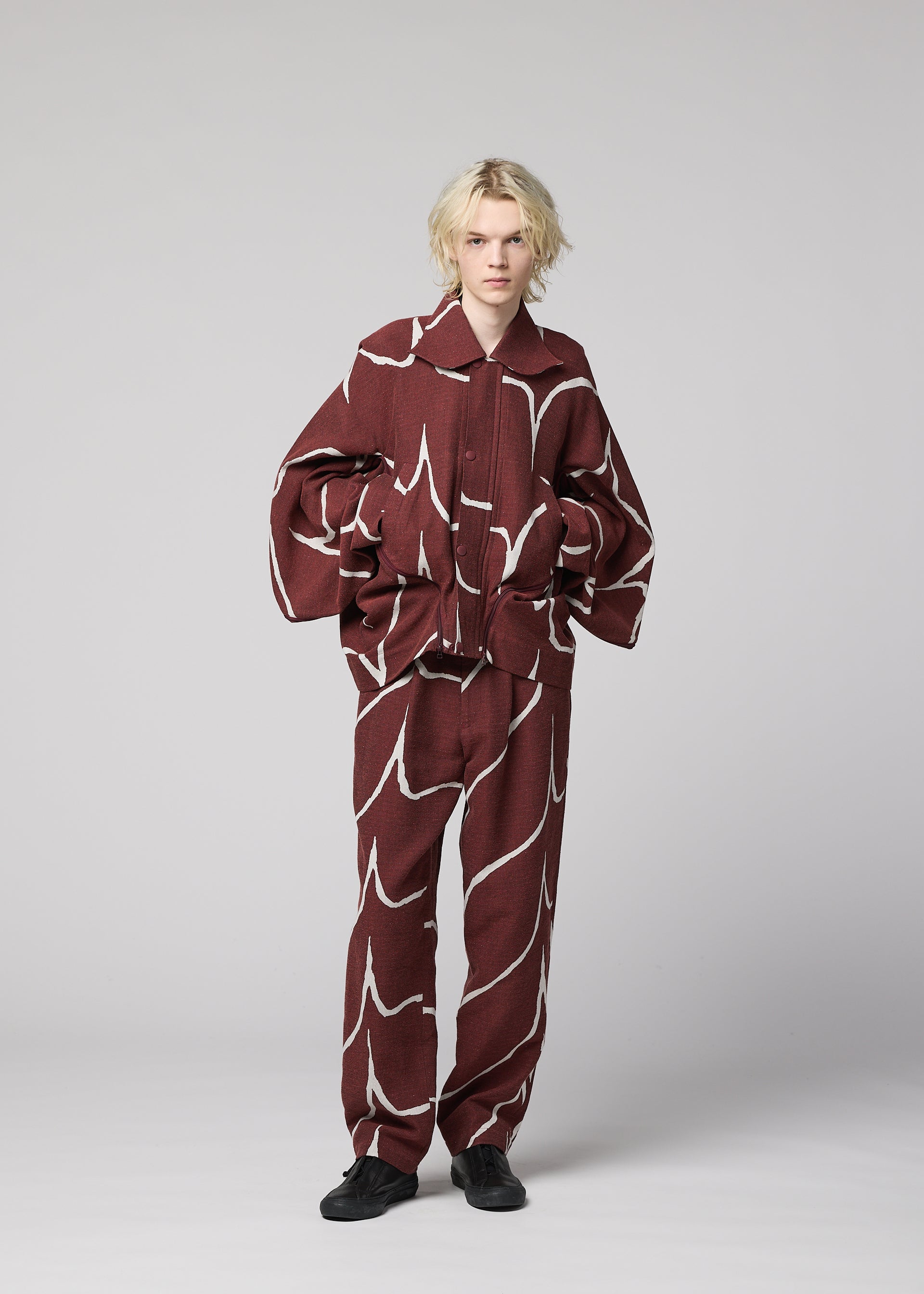 Zoomed view A person with blonde hair stands against a plain background, wearing the IM MEN EARTH matching maroon jacket and pants set with bold white wavy lines, paired with black shoes.
