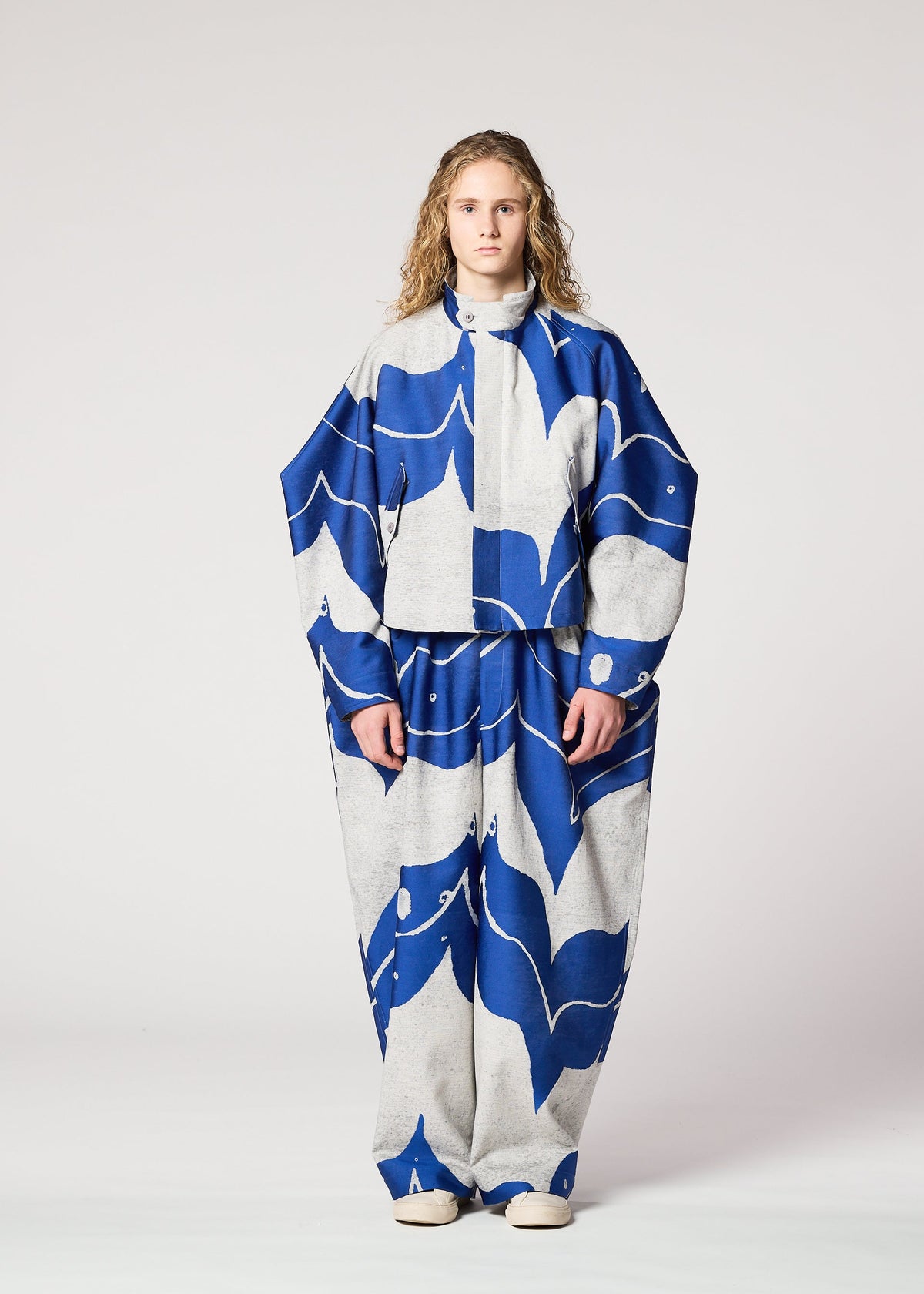 A person with long curly hair models the IM MEN INLAY suit—loose, oversized, and crafted from recycled polyester with bold blue and white abstract patterns on a gray background—against a plain white backdrop. view 5 of 8