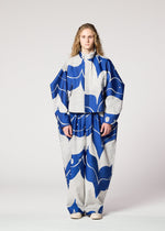 A person with long curly hair models the IM MEN INLAY suit—loose, oversized, and crafted from recycled polyester with bold blue and white abstract patterns on a gray background—against a plain white backdrop.