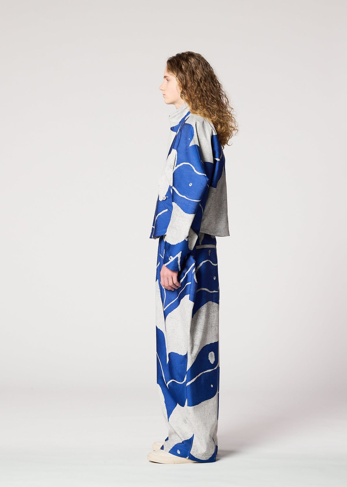 A person with long curly hair stands in profile, wearing the INLAY jacket and wide-legged pants by IM MEN, featuring a blue and gray abstract ceramic-inspired pattern in recycled polyester, set against a plain light background. view 6 of 8