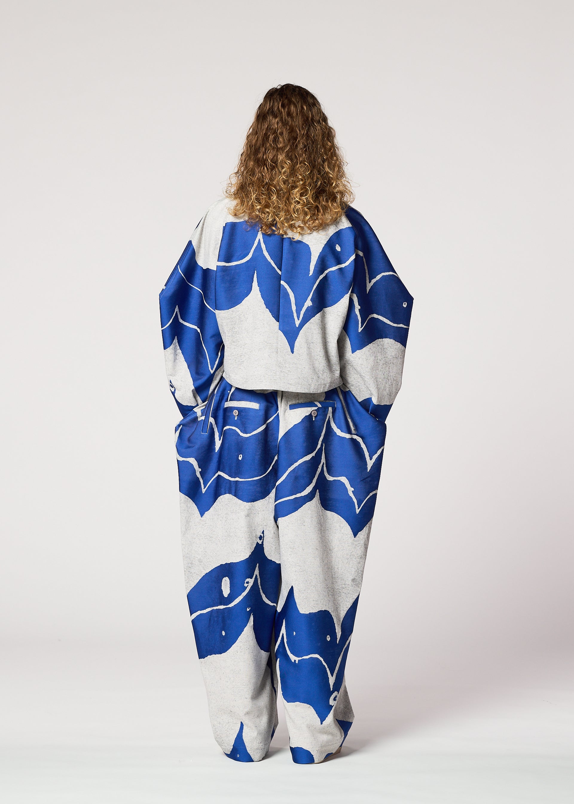 Zoomed view A person with curly hair stands facing away, wearing the INLAY jacket and wide-legged pants by IM MEN, featuring a bold blue and white wave pattern. The abstract set is crafted from recycled polyester on a jacquard loom.