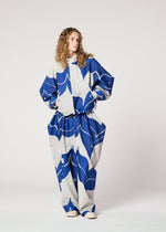 A person with long curly hair stands against a plain background, wearing IM MEN’s INLAY—a loose-fitting recycled polyester outfit featuring a bold blue and gray abstract pattern. Their hands are in their pockets as they look to the side.