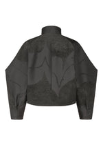 The image shows the back view of the INLAY jacket by IM MEN: a dark gray, long-sleeve, high-neck piece with a ceramic-inspired abstract pattern in lighter gray. The boxy cut highlights its jacquard-woven textured fabric.