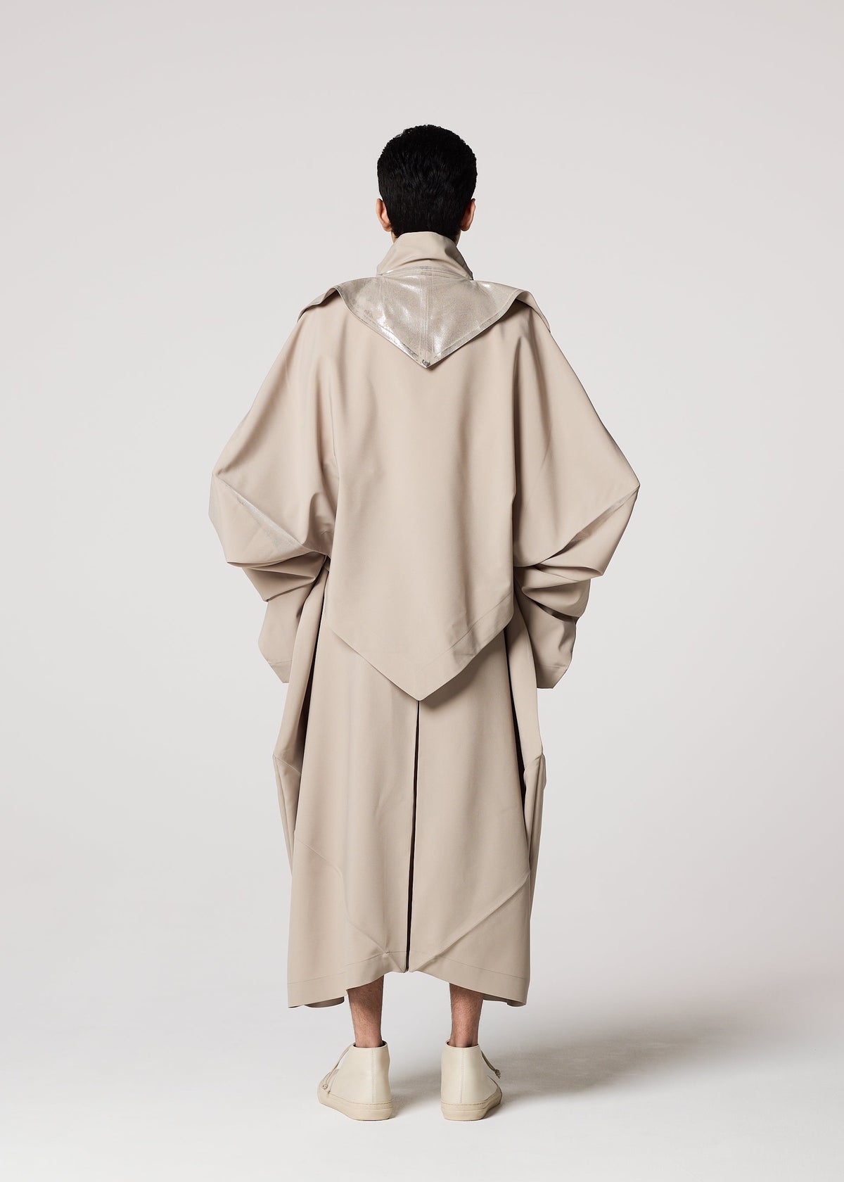 A person faces away, wearing a Shoji Kamoda-inspired long beige coat with wide sleeves and a shiny silver collar, paired with IM MEN's GINTO FLAT light-colored shoes against a plain background. view 8 of 9