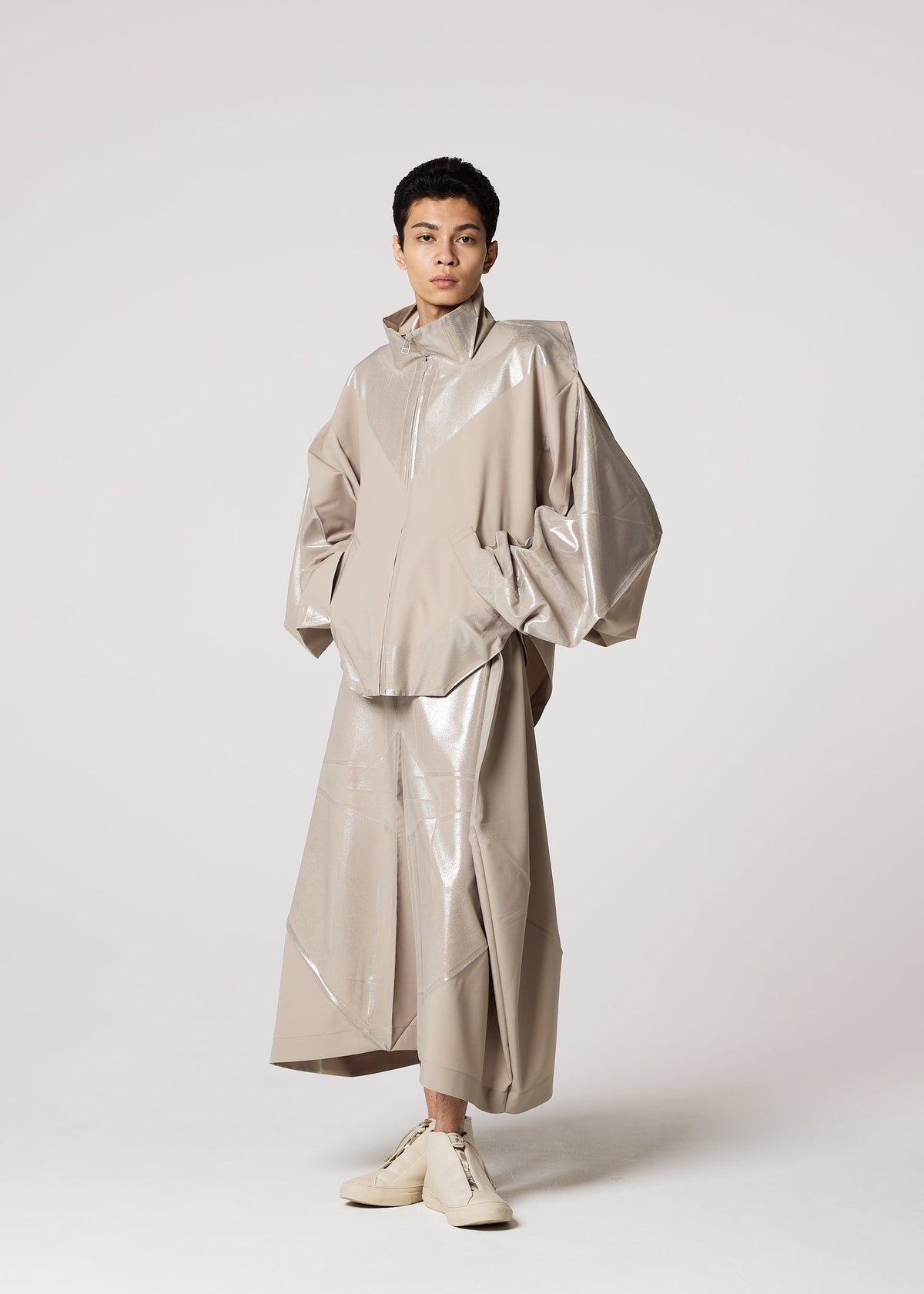 A person with short dark hair wears IM MEN’s shiny, loose-fitting plant-based polyester jacket and matching long skirt, inspired by Shoji Kamoda, paired with beige GINTO FLAT ankle-high shoes, against a plain backdrop. view 9 of 9