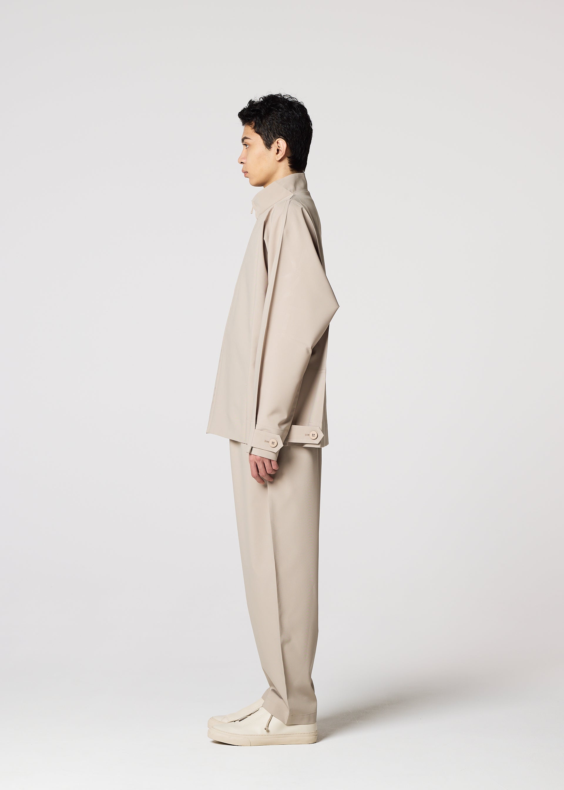 Zoomed view A person with short dark hair stands in profile against a white background, wearing IM MEN's FLAT matching beige long-sleeve top and loose pants, light shoes, and carrying a compact portable jacket over one arm.