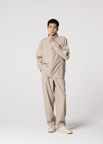 A person with short dark hair and a neutral expression wears the IM MEN FLAT set: a loose beige long-sleeve shirt, matching wide-leg trousers, a compact jacket, and white sneakers against a light background.