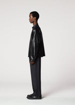 A person in profile against a plain background, wearing the IM MEN GINTO black plant-derived artificial suede leather-textured jacket, paired with dark pleated pants and black shoes.