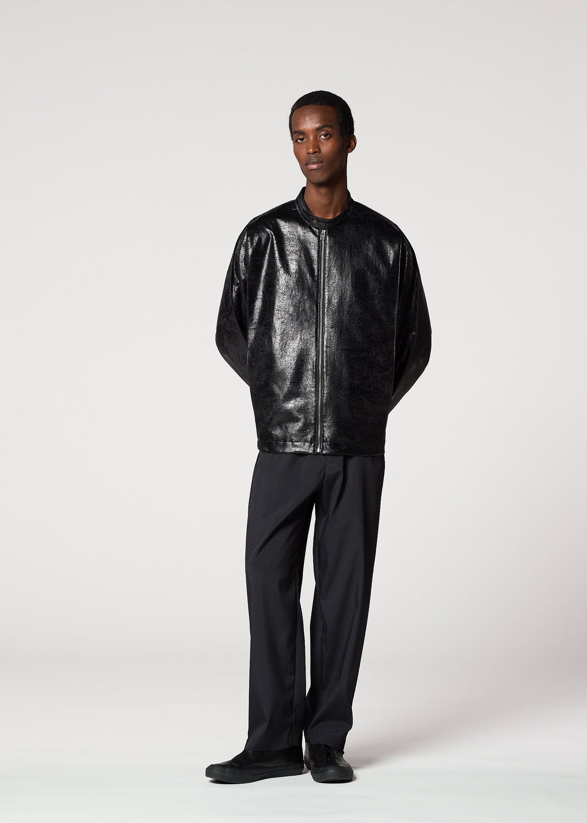 A person stands against a plain light background wearing the GINTO shiny silver foil jacket by IM MEN, paired with loose black trousers and black shoes, hands behind their back. view 5 of 5
