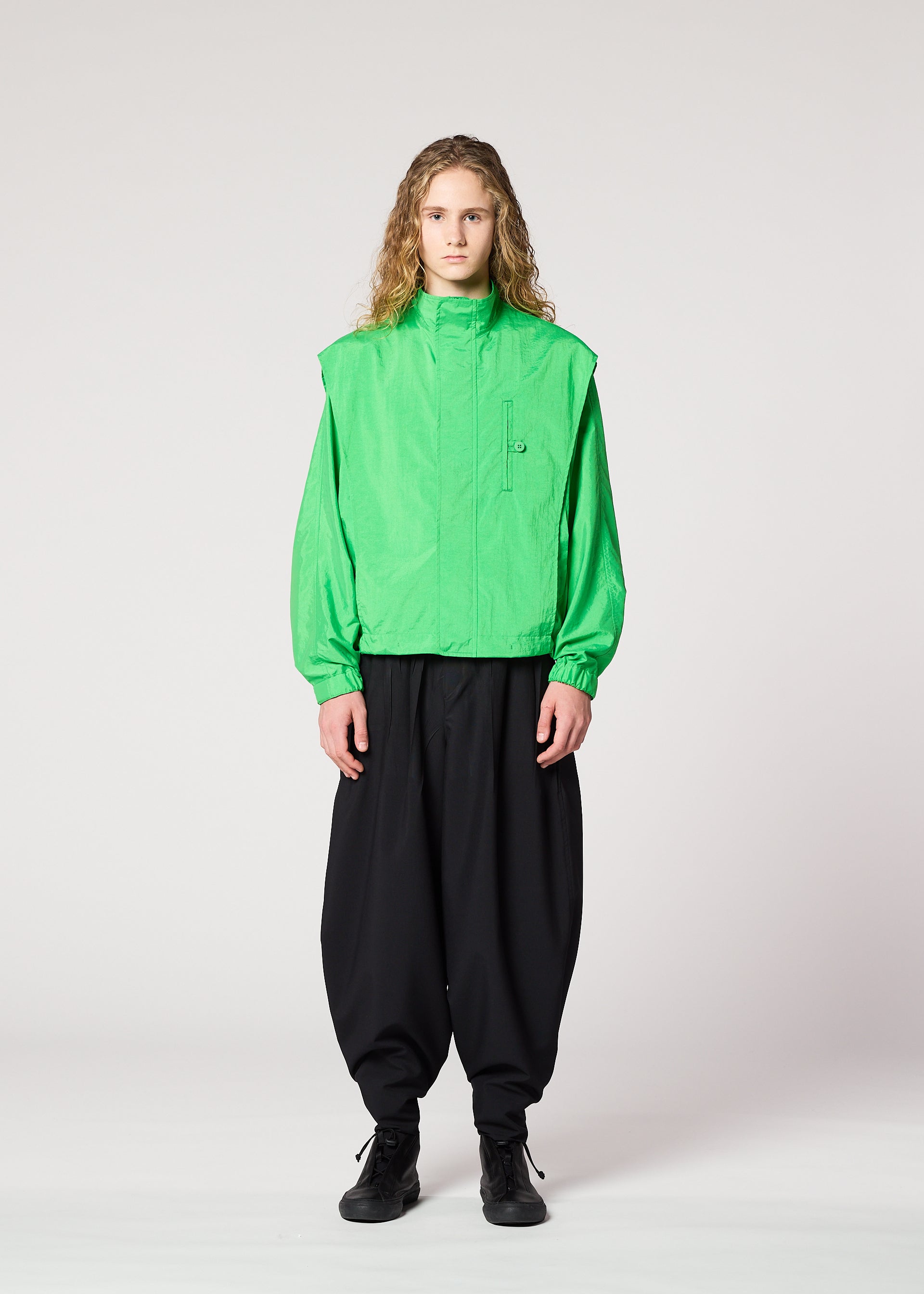 Zoomed view A person with long curly hair faces forward, wearing the IM MEN FISHING NET NYLON bright green jacket made from recycled fishing nets, paired with loose black pants and black shoes, against a plain light background.