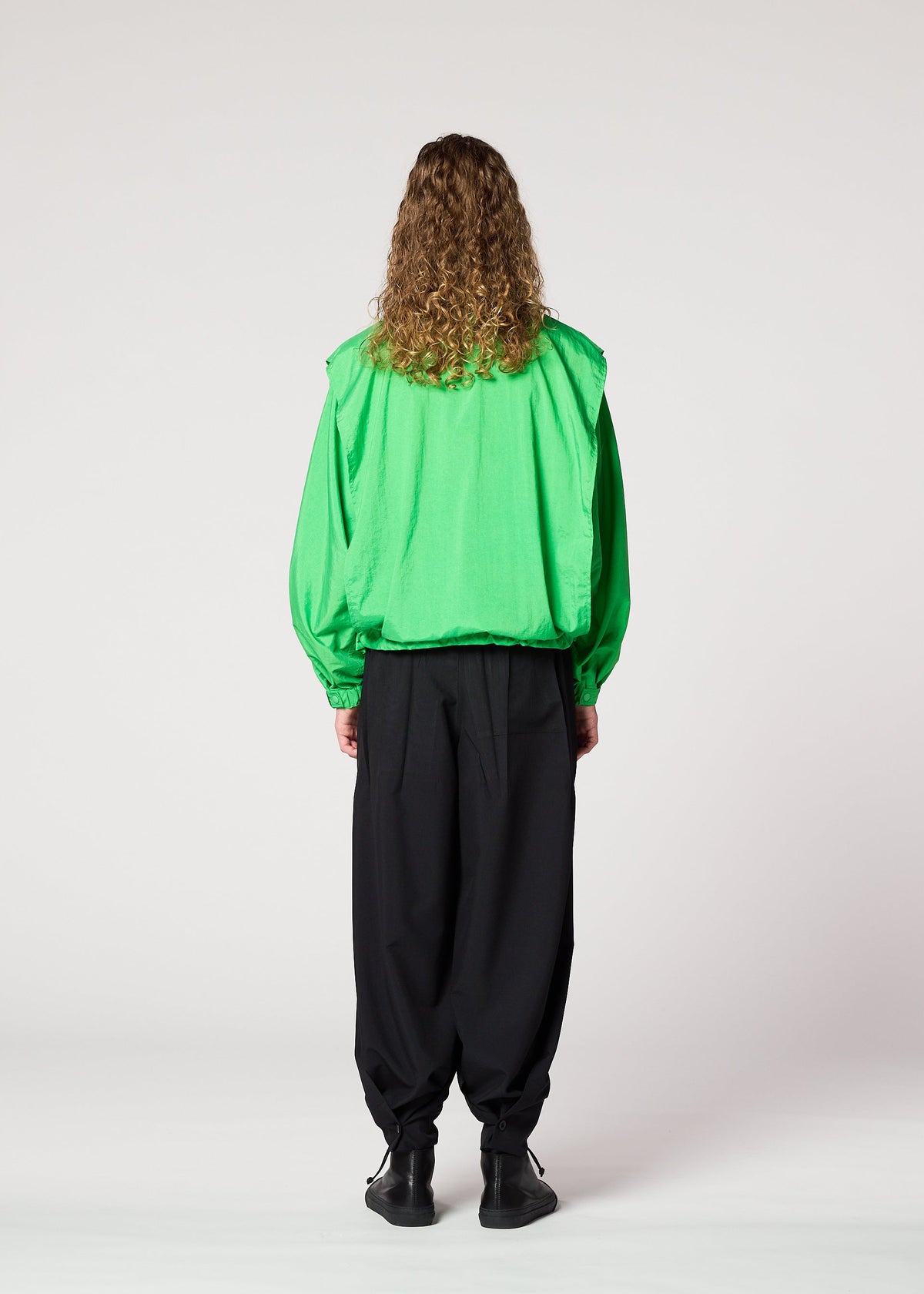 A person with long curly hair stands facing away, wearing the IM MEN FISHING NET NYLON bright green jacket made from recycled fishing nets, paired with loose black pants and black shoes, against a plain white background. view 8 of 9