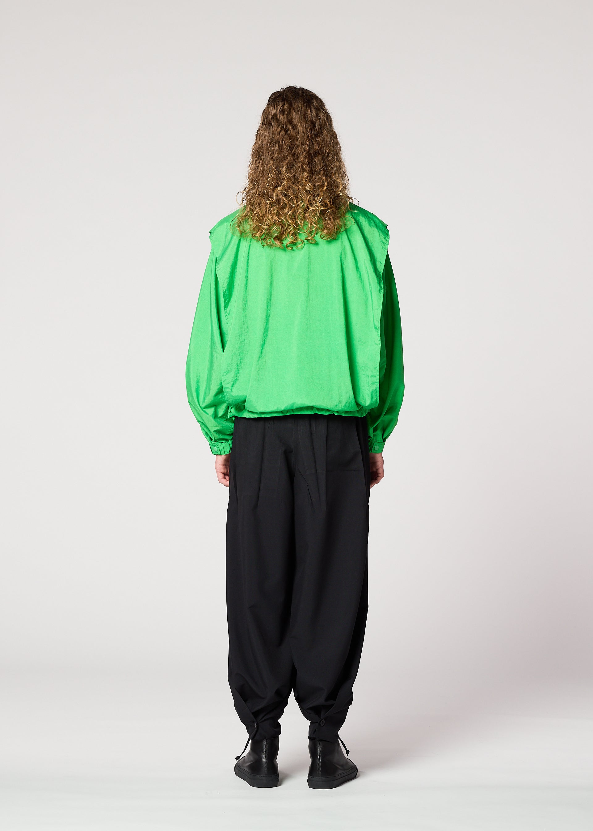 Zoomed view A person with long curly hair stands facing away, wearing the IM MEN FISHING NET NYLON bright green jacket made from recycled fishing nets, paired with loose black pants and black shoes, against a plain white background.