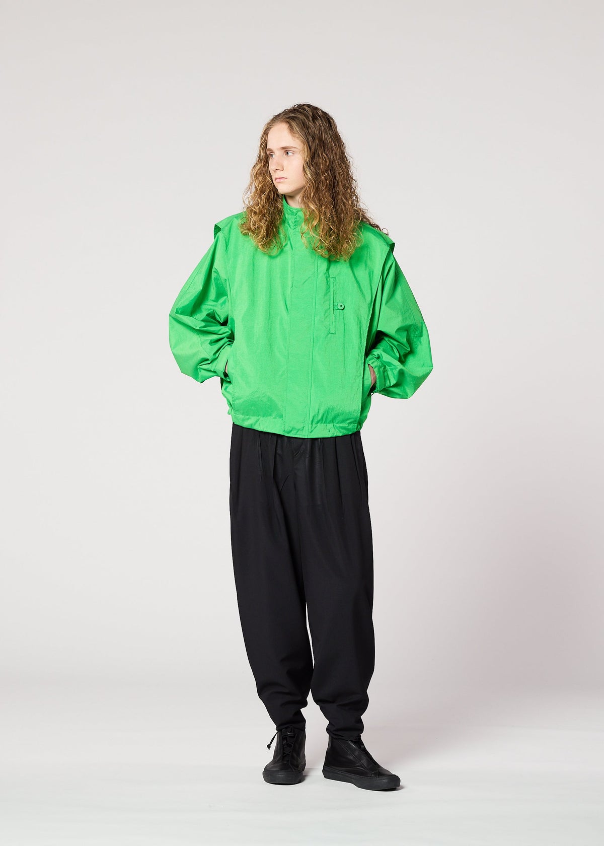 A person with long curly hair stands against a plain background, wearing the IM MEN FISHING NET NYLON bright green oversized jacket made from recycled fishing nets, loose black pants, and black shoes, looking to the side with hands in pockets. view 9 of 9