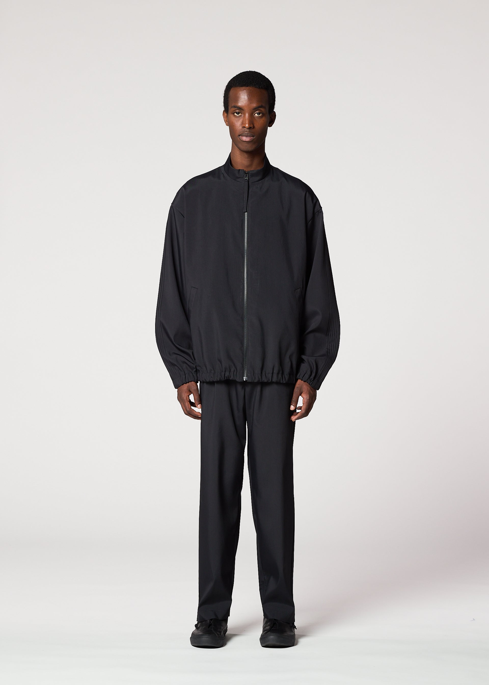 Zoomed view A man stands facing forward against a plain light background, wearing the IM MEN AIR PLEATS wrinkle-resistant zip-up black jacket and matching trousers. His arms are by his sides and his expression is neutral.