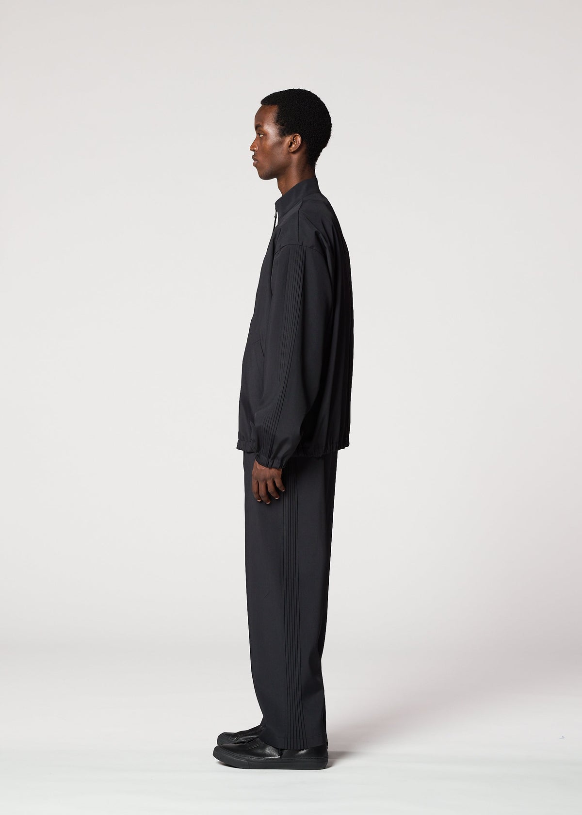 A man stands in profile against a plain white background, dressed in IM MEN's AIR PLEATS lightweight, wrinkle-resistant black jacket and matching pants, paired with black shoes. view 7 of 8