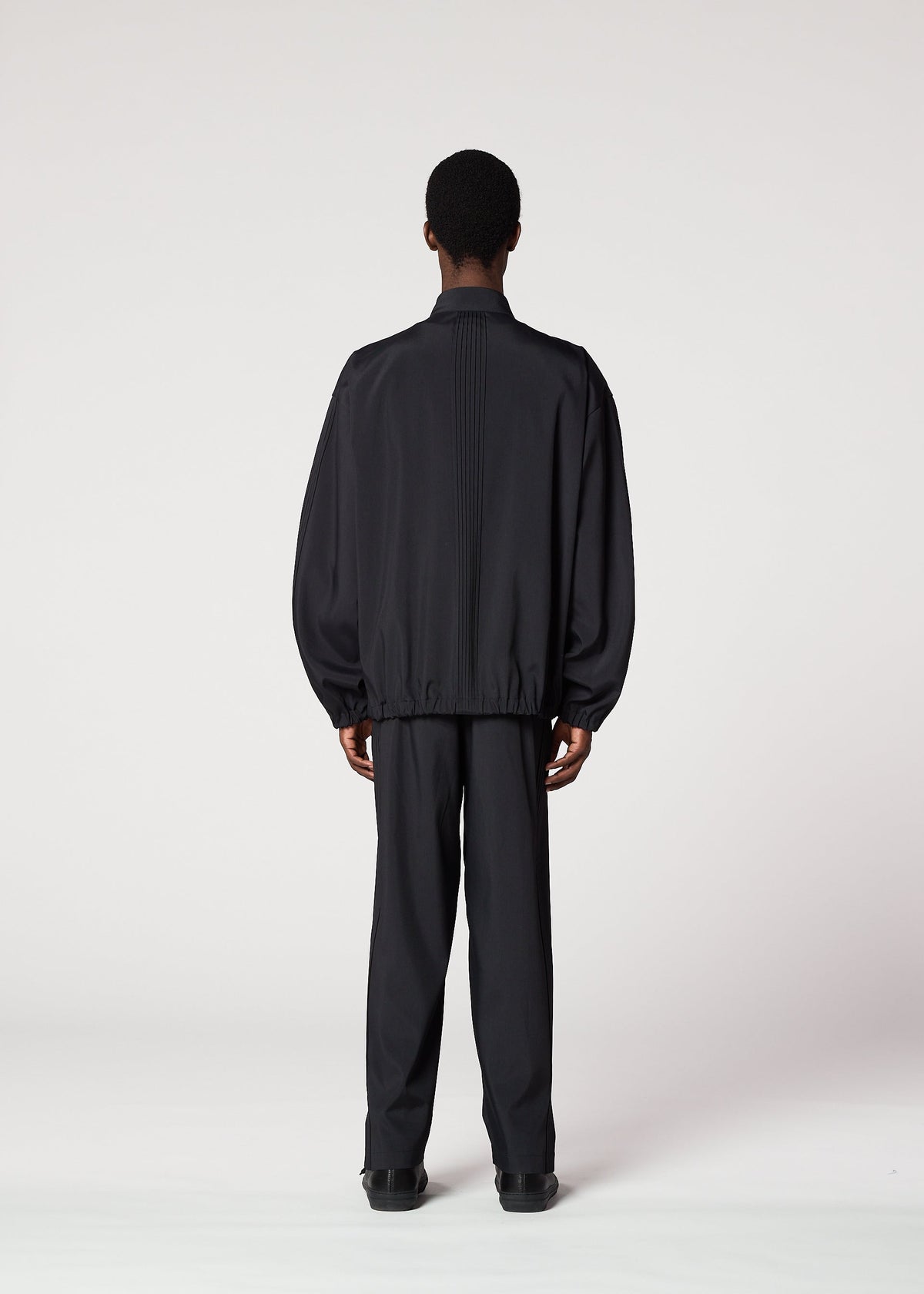 A person with short hair faces away from the camera, wearing IM MEN’s AIR PLEATS lightweight black jacket and matching pants designed for freedom of movement, set against a plain white background. view 8 of 8