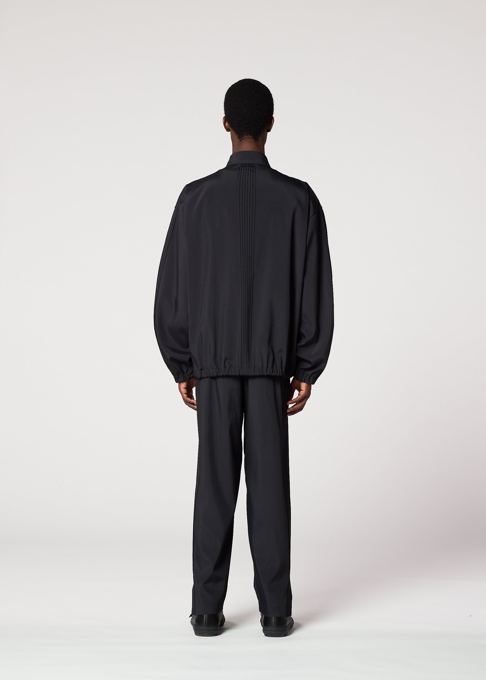 Zoomed view A person with short hair faces away from the camera, wearing IM MEN’s AIR PLEATS lightweight black jacket and matching pants designed for freedom of movement, set against a plain white background.