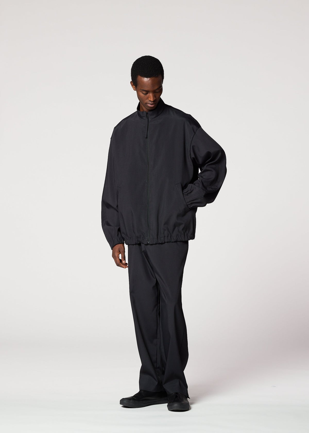 A person stands against a plain background wearing the IM MEN AIR PLEATS set—a loose-fitting, wrinkle-resistant zip-up jacket and matching pants with black shoes—offering freedom of movement as they look slightly downward with one hand in their pocket. view 5 of 8