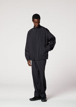 A person stands against a plain background wearing the IM MEN AIR PLEATS set—a loose-fitting, wrinkle-resistant zip-up jacket and matching pants with black shoes—offering freedom of movement as they look slightly downward with one hand in their pocket.