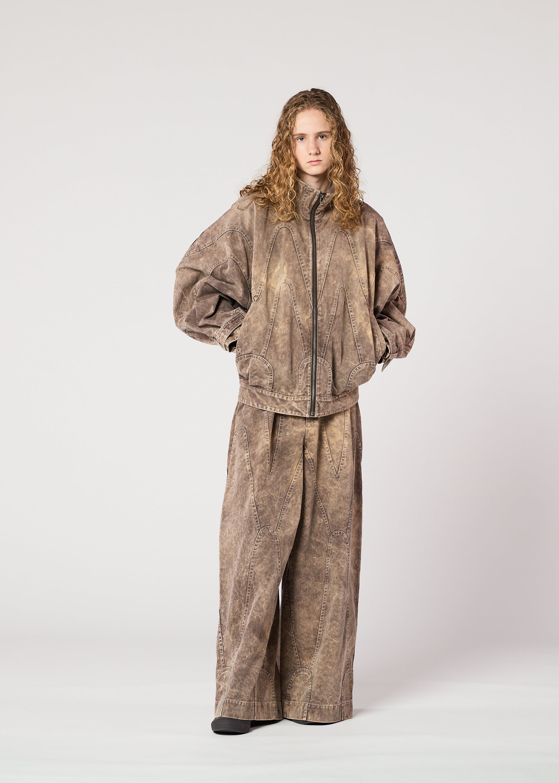 Zoomed view A young person with curly hair models the IM MEN ELEMENTS collection: a loose brown ceramic artist jacket and matching wide-leg pants with a subtle color-fading effect, posing against a plain white background.