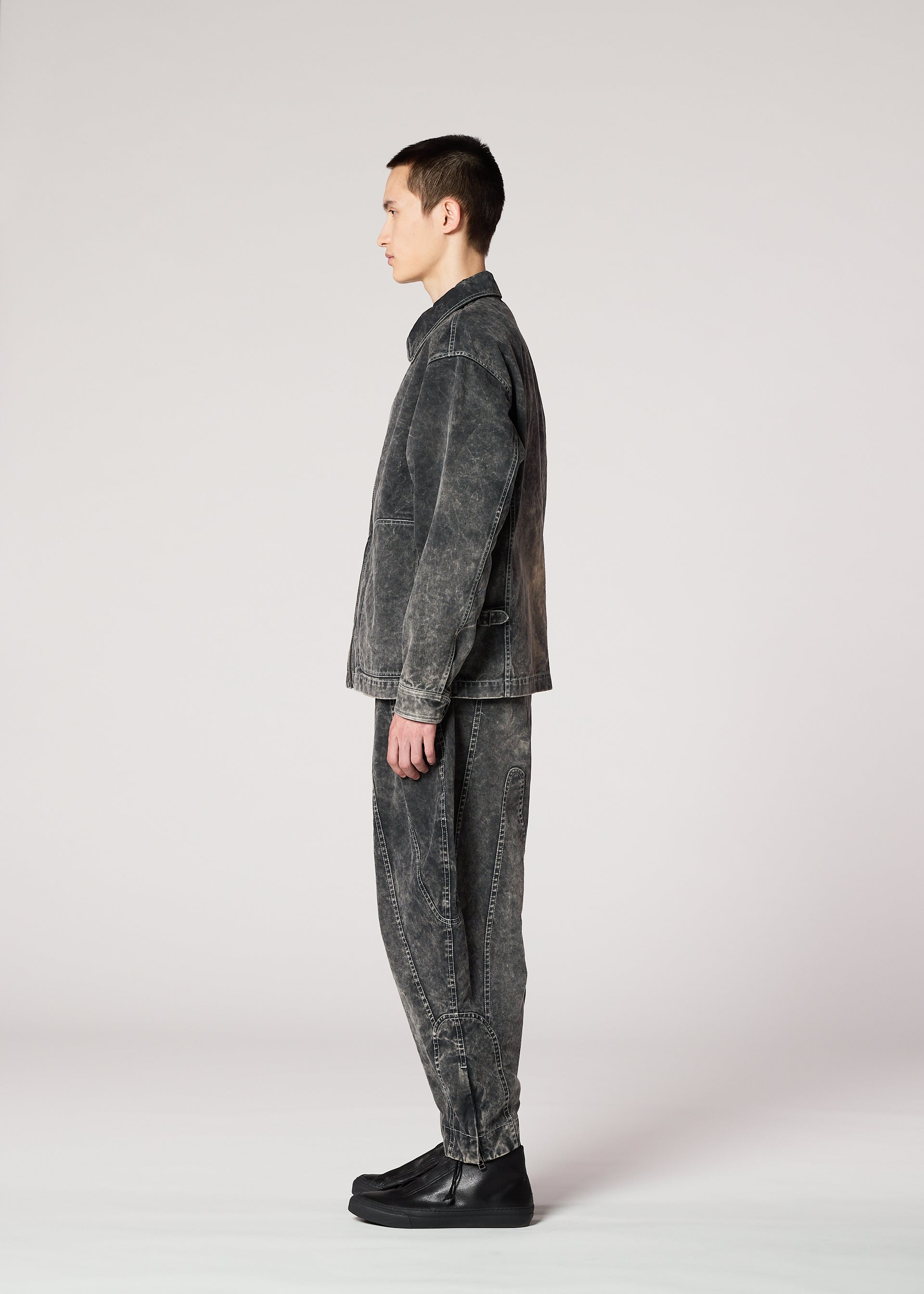 Zoomed view A person stands in profile against a plain light background, wearing IM MEN’s ELEMENTS set: a matching faded black denim jacket and pants with black shoes, featuring color-fading inspired by ceramic artist Shoji Kamoda’s signature style.
