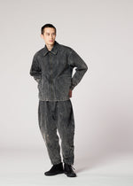 Against a plain light background, a person wears the IM MEN ELEMENTS faded black denim jacket and pants set, featuring a subtle color-fading effect inspired by Shoji Kamoda’s textured ceramics. They look to the side with hands behind their back.