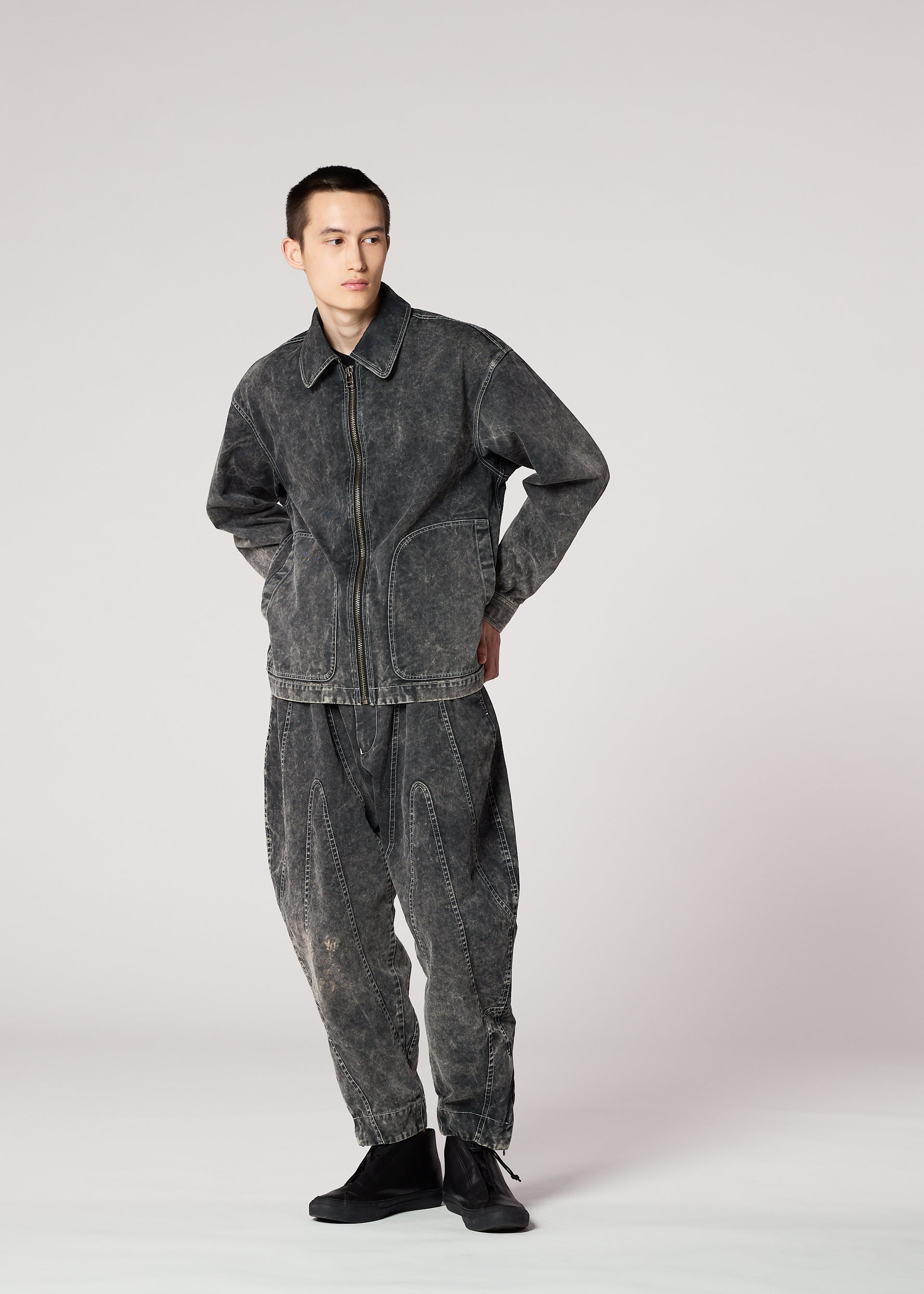 Zoomed view Against a plain light background, a person wears the IM MEN ELEMENTS faded black denim jacket and pants set, featuring a subtle color-fading effect inspired by Shoji Kamoda’s textured ceramics. They look to the side with hands behind their back.