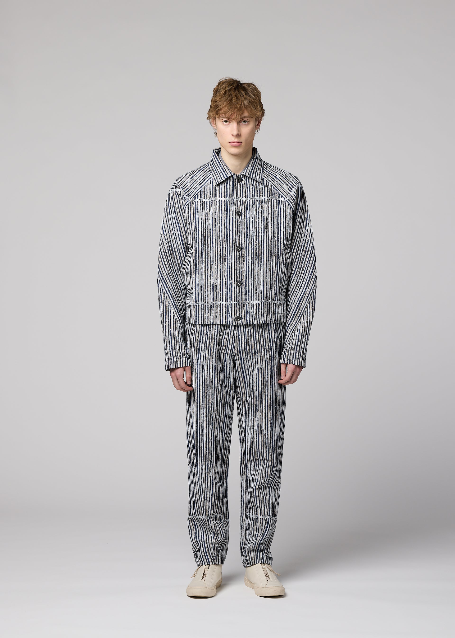 Zoomed view A person in the IM MEN CURVINESS set—cotton linen jacket and pants with vertical black and white stripes—stands against a light gray background in cream shoes, looking straight ahead with a neutral expression.
