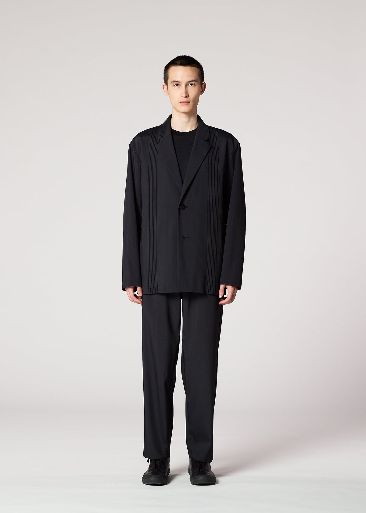 A person stands facing forward against a plain light background, wearing IM MEN's AIR PLEATS BLACK—a loose-fitting, wrinkle-resistant suit—paired with a black shirt and black shoes, hands relaxed by their sides. view 4 of 7
