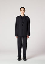 A person stands facing forward against a plain light background, wearing IM MEN's AIR PLEATS BLACK—a loose-fitting, wrinkle-resistant suit—paired with a black shirt and black shoes, hands relaxed by their sides.