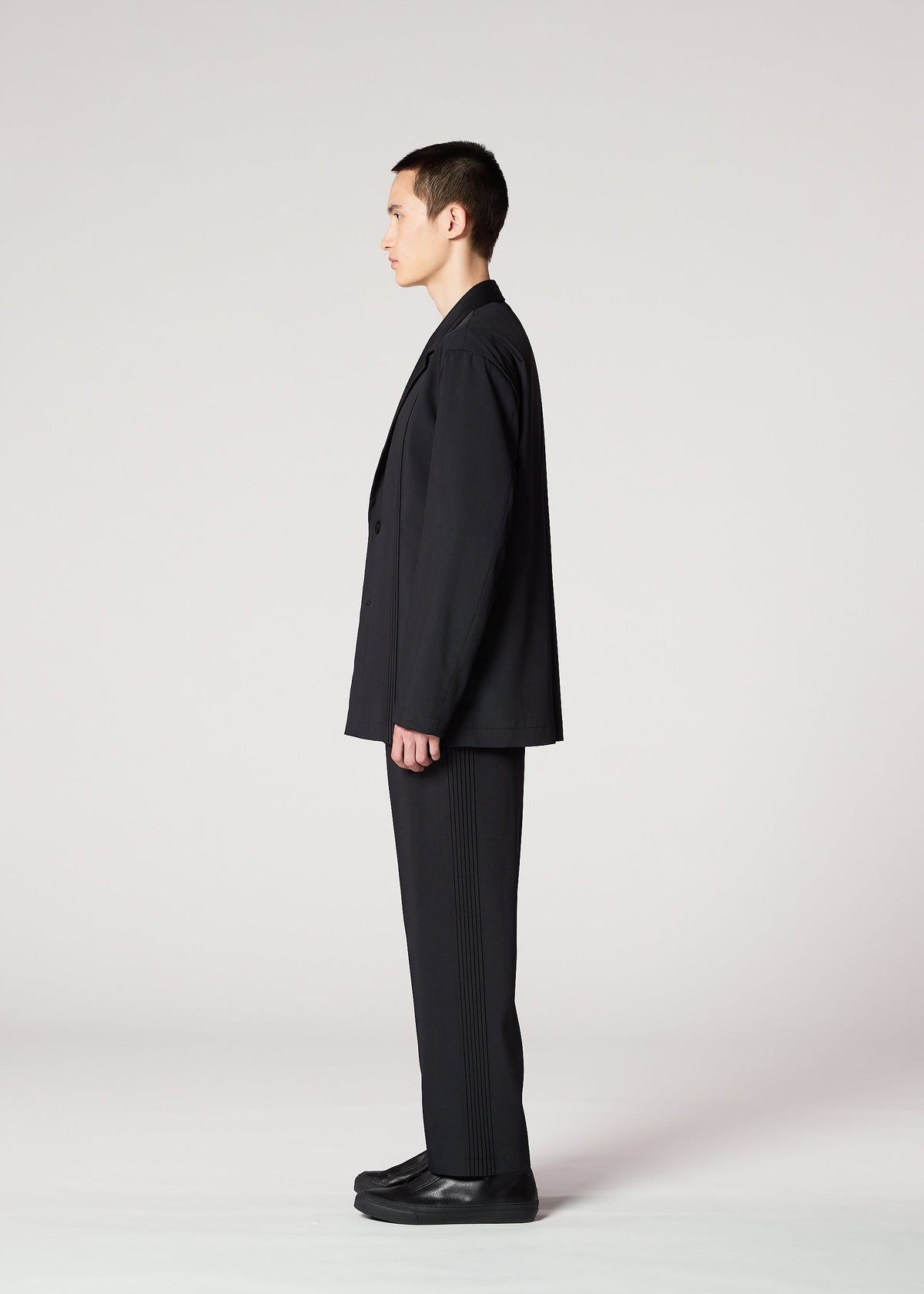 A person with short dark hair wears IM MEN's AIR PLEATS BLACK suit and black shoes, standing sideways facing left against a plain light gray background. view 6 of 7