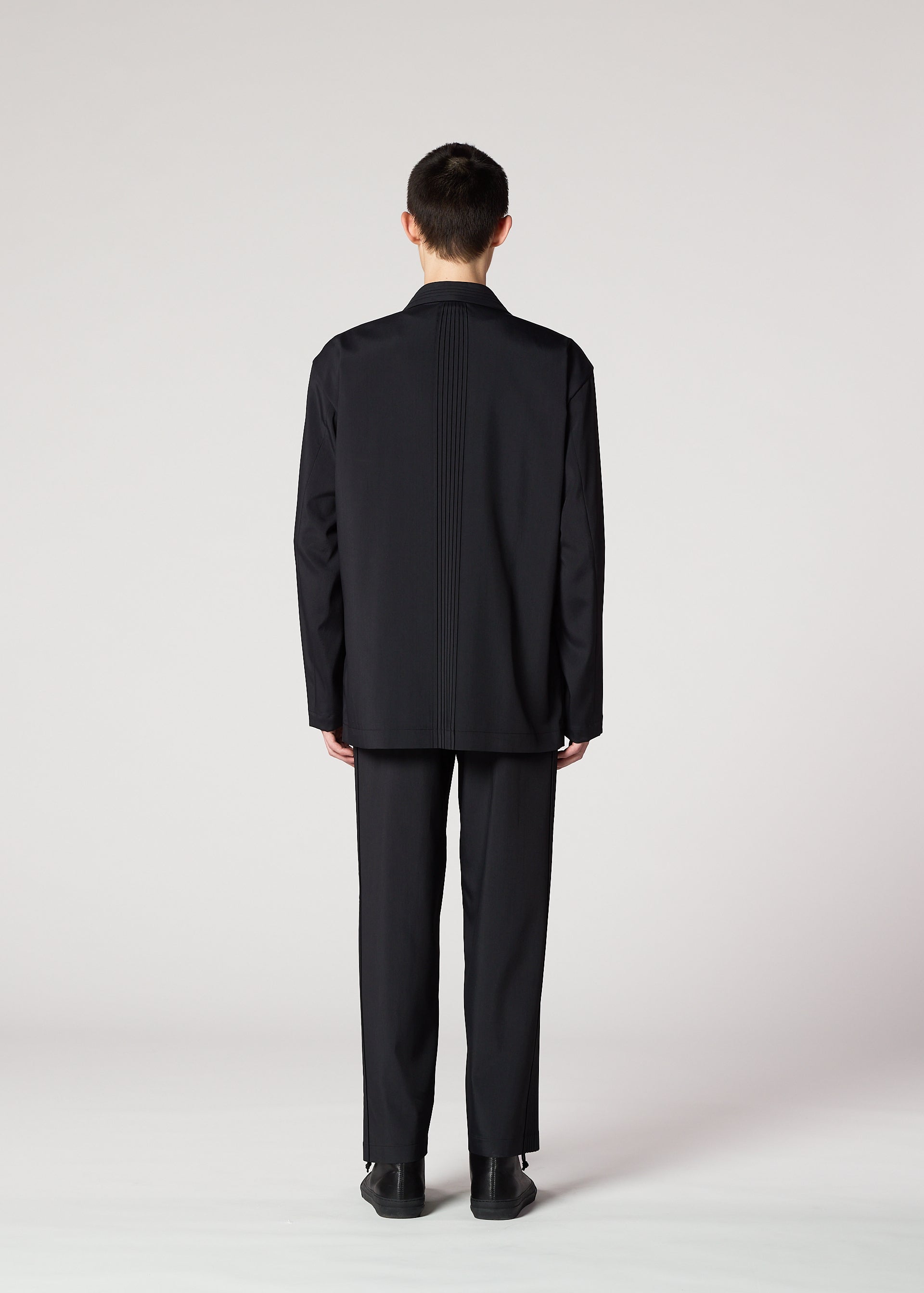 Zoomed view A person with short dark hair stands facing away, wearing the wrinkle-resistant IM MEN AIR PLEATS BLACK tailored jacket and pants. The background is plain and light, and their hands rest naturally by their sides.