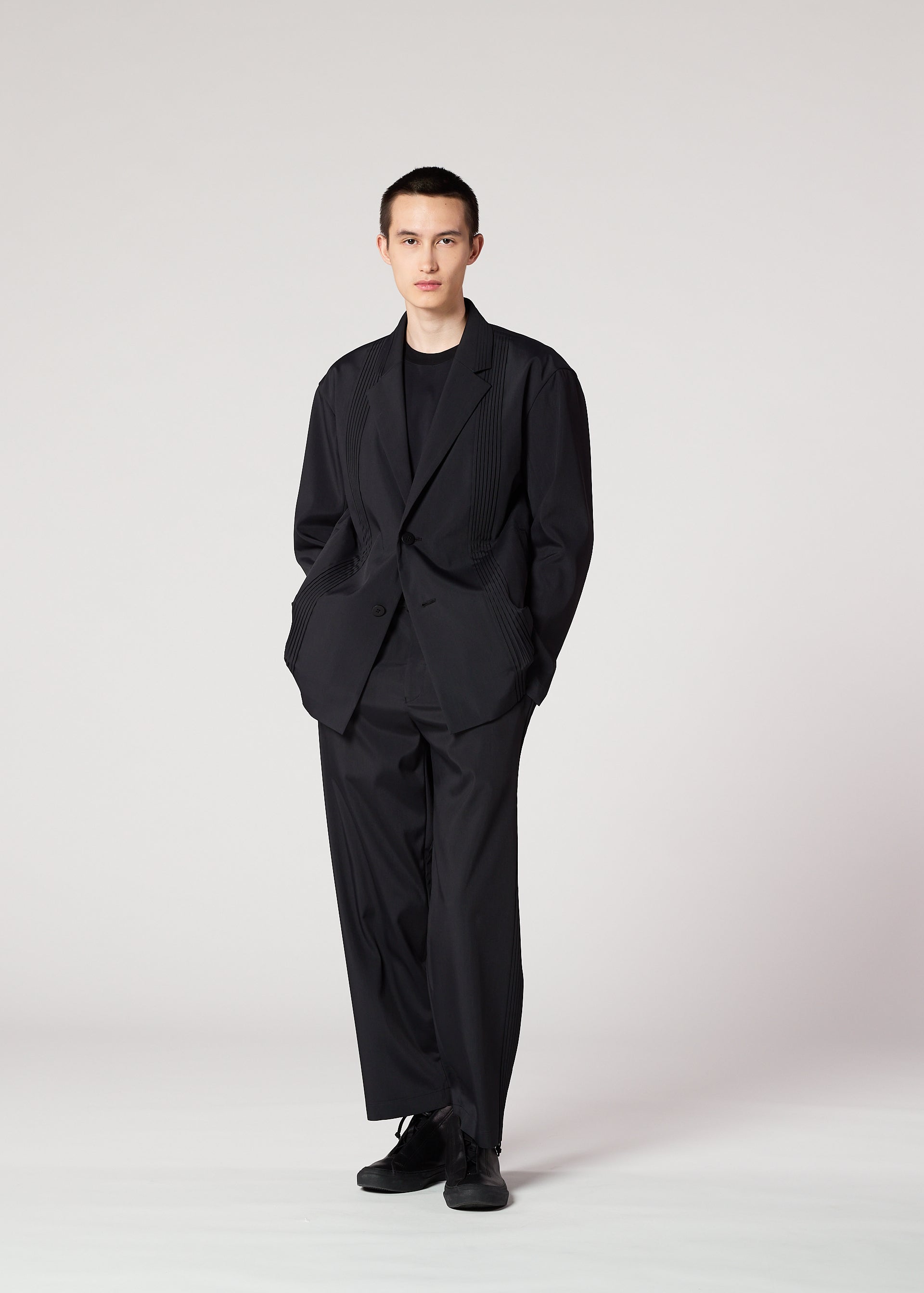 Zoomed view A person stands against a plain light background, wearing the IM MEN AIR PLEATS BLACK jacket with matching shirt and shoes, hands in pockets, looking directly at the camera with a neutral expression.