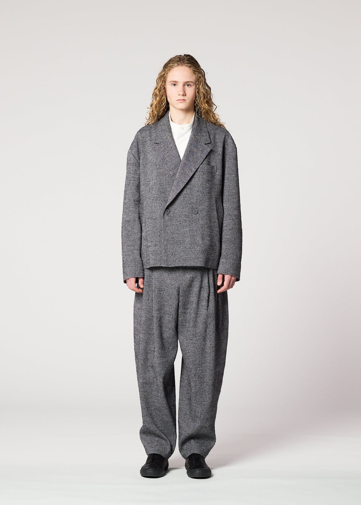 A person with long curly hair faces forward, wearing the IM MEN TEXTURES loose gray suit with a double-breasted jacket and wide pants featuring ceramic-inspired textile details, a white shirt underneath, and black shoes against a plain light background. view 5 of 8
