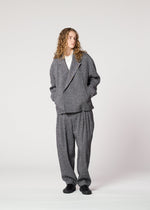 A person with long curly hair stands hands in pockets, wearing the IM MEN TEXTURES ceramic-inspired double-breasted gray suit over a white shirt and black shoes against a plain light background.