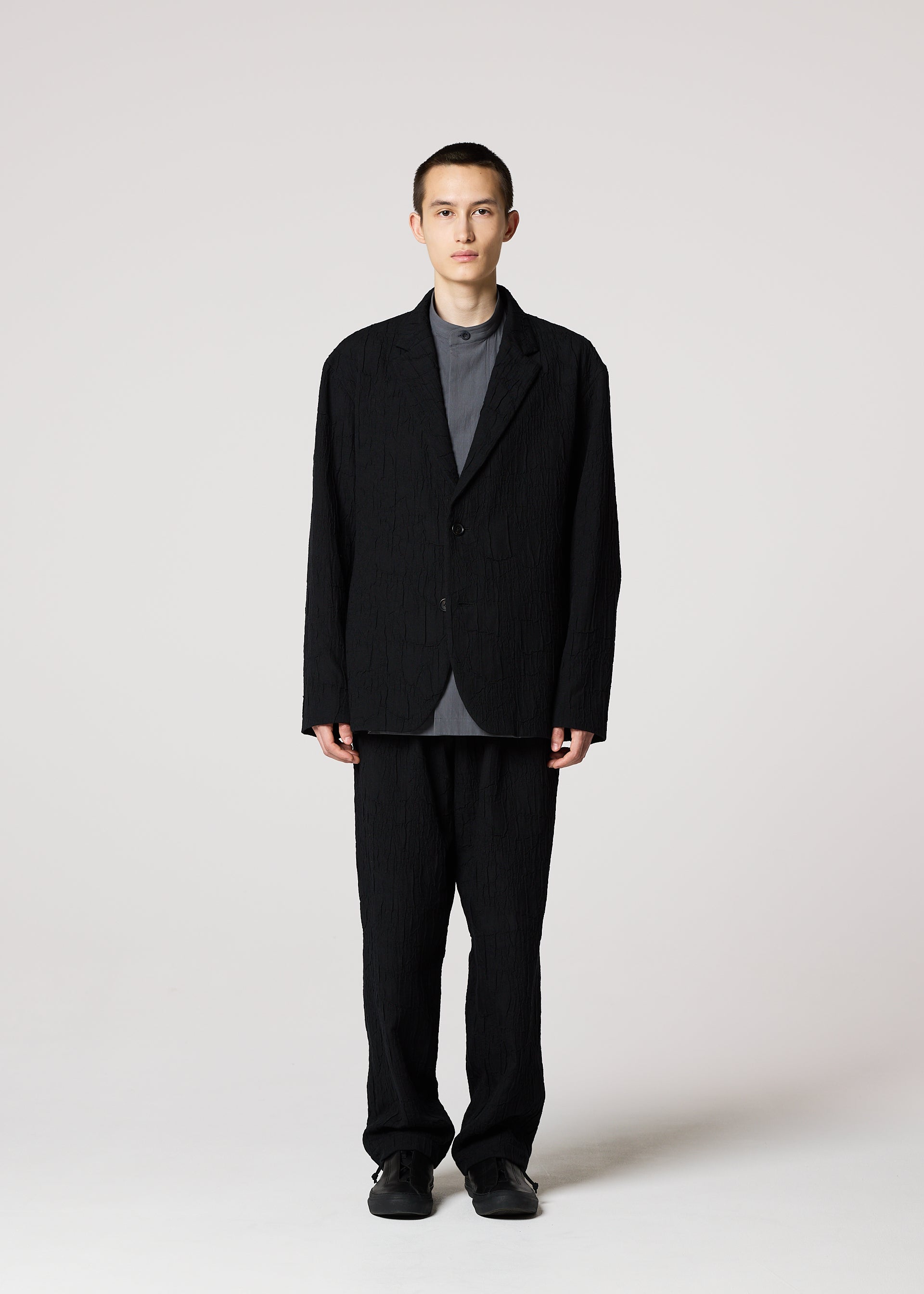 Zoomed view A person stands facing forward, wearing the IM MEN SHRINK JACQUARD tailored jacket and matching pants with a subtle wood-grain pattern over a gray shirt and black shoes, set against a plain light background.