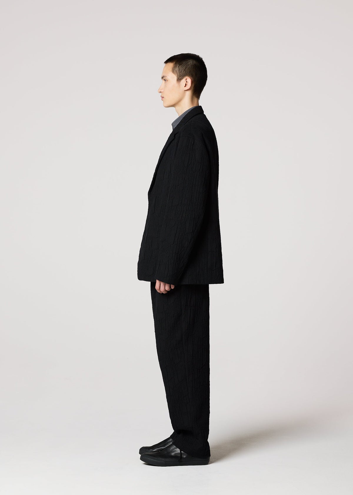 A man stands in profile against a plain background, wearing the IM MEN SHRINK JACQUARD tailored jacket with a subtle wood-grain woven pattern, paired with a dark shirt and black shoes. His hands rest at his sides as he looks straight ahead. view 6 of 8