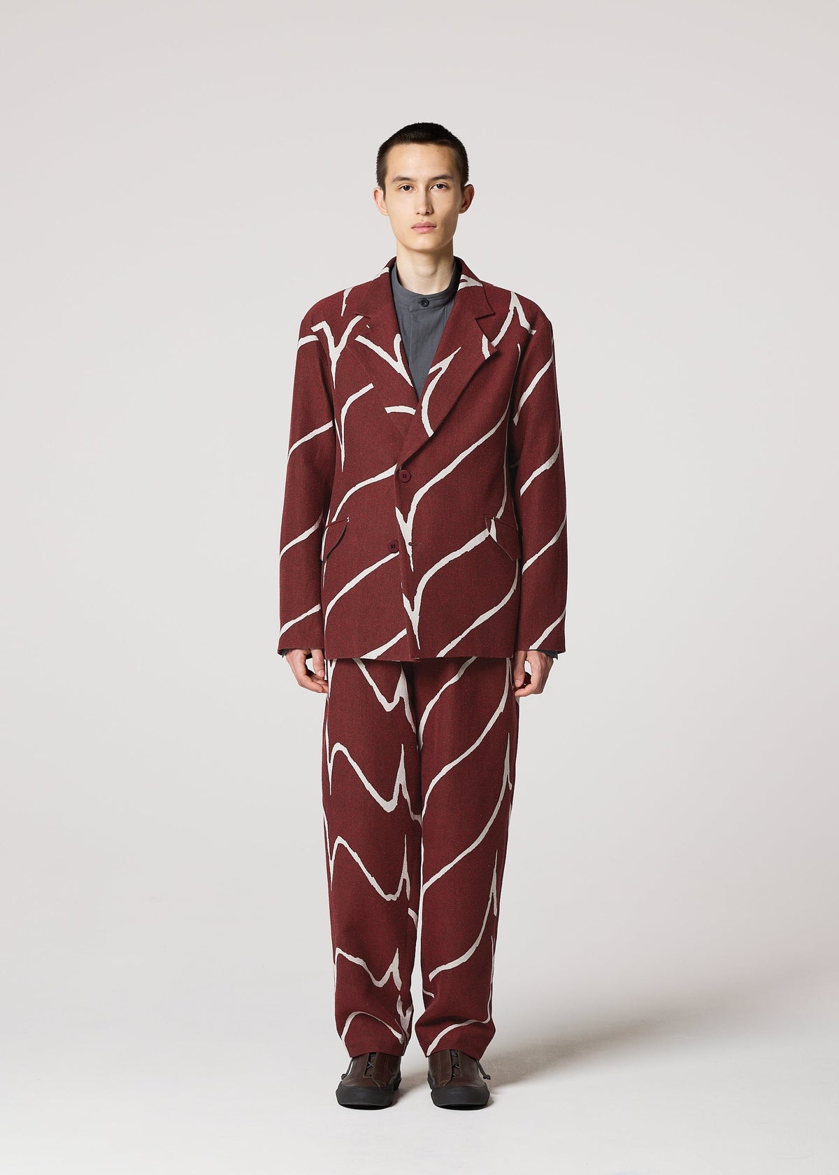 A person faces forward wearing the IM MEN EARTH maroon jacquard wave-pattern jacket over a dark shirt with dark shoes, set against a plain light background. view 5 of 9