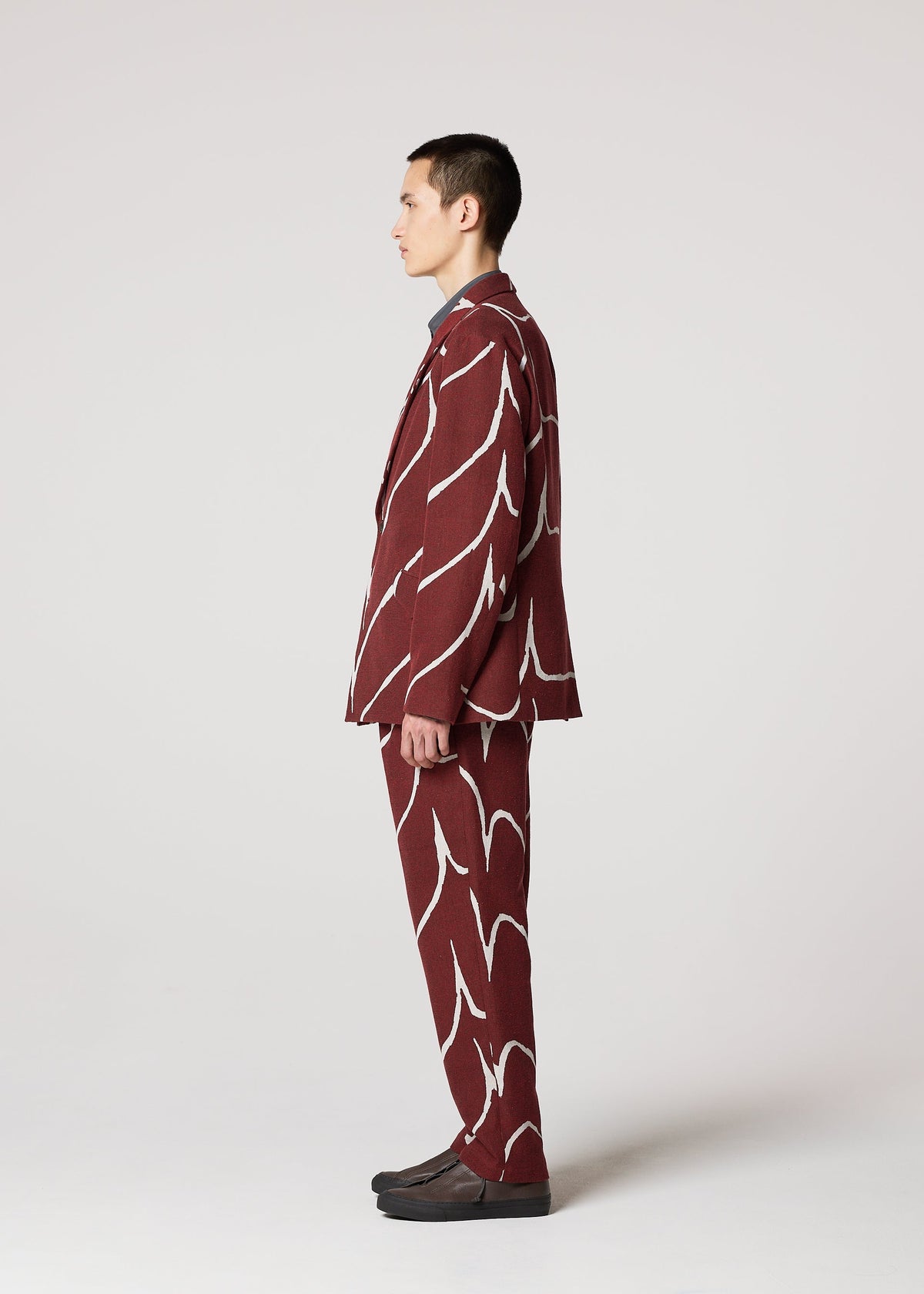 A person stands in profile against a plain background, wearing IM MEN’s EARTH tailored maroon suit featuring a bold jacquard wave pattern, paired with dark shoes. view 6 of 9